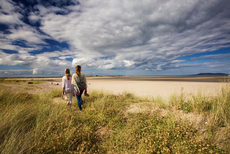 Two women walking along the sandy shore of Malahide Beach, a popular coastal day trip from The Trinity City Hotel, Dublin.